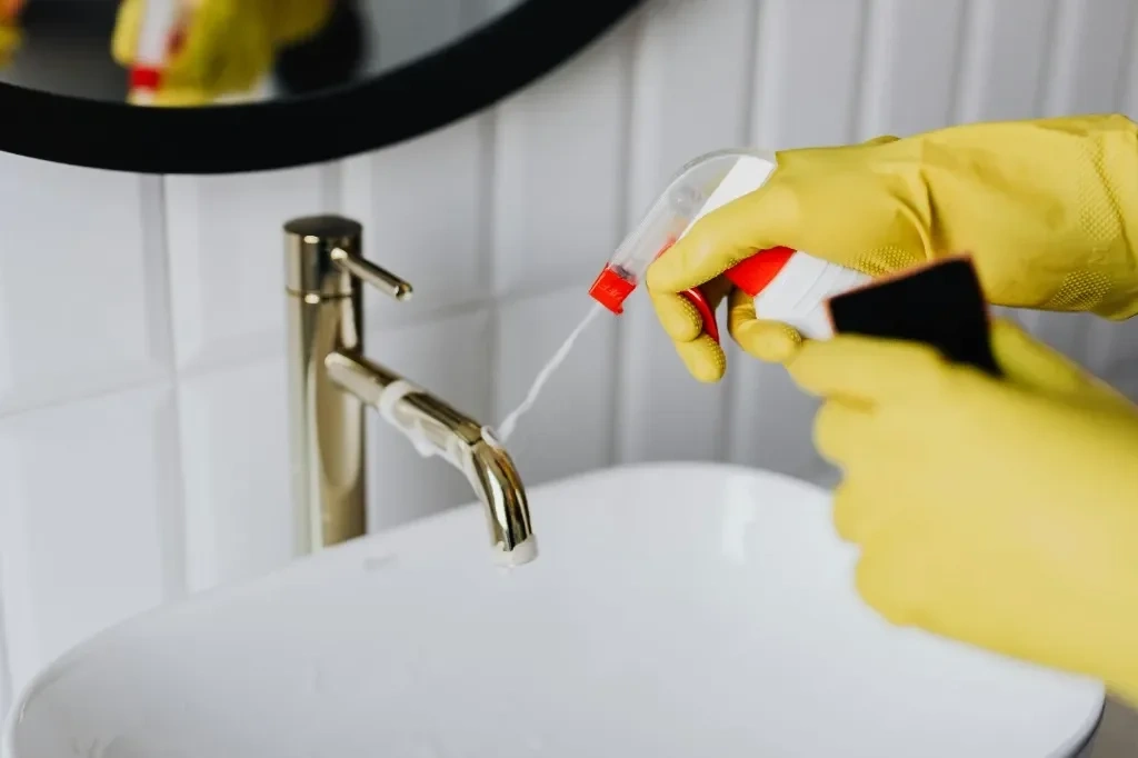 Professional housekeeper in yellow rubber gloves spraying eco-friendly cleaning sanitizer on a luxury gold faucet and sink