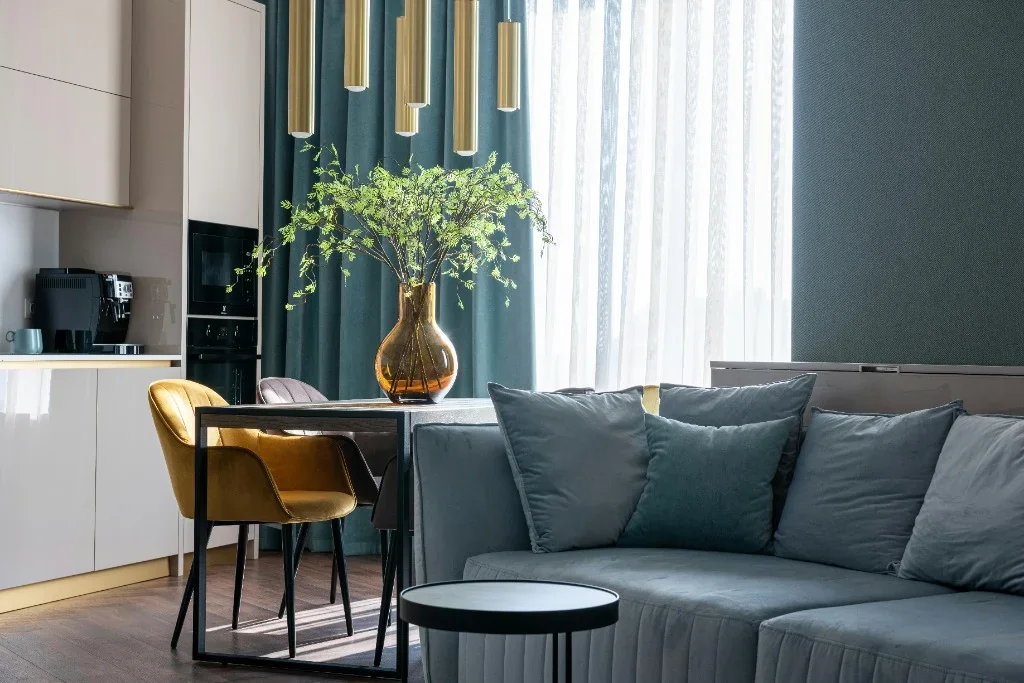 An immaculately clean and luxurious open-concept Toronto apartment featuring a plush grey sofa, a stylish dining table with a yellow velvet chair, and a decorative amber vase with fresh green branches in a sunlit room.
